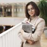 Education, business and women concept. Portrait of young attractive, elegant female tutor, young teacher or student carry studying books and laptop, standing in hall smiling camera